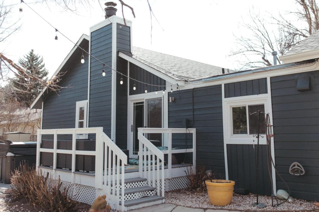 A modern house with a dark blue exterior and white trim, featuring a porch with a railing, large windows, and string lights. The porch leads to a backyard with decorative stones and a yellow planter.