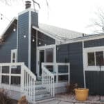 A modern house with a dark blue exterior and white trim, featuring a porch with a railing, large windows, and string lights. The porch leads to a backyard with decorative stones and a yellow planter.