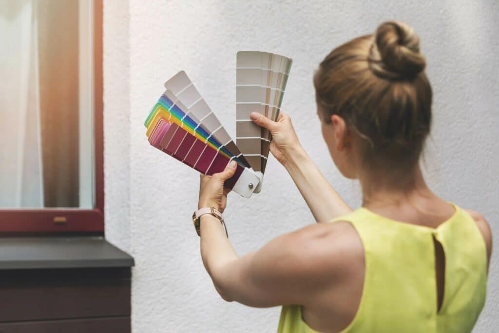 A woman holding multiple color swatches in front of a window, analyzing shades for a design project. She has long hair styled in a bun and is wearing a sleeveless yellow top.