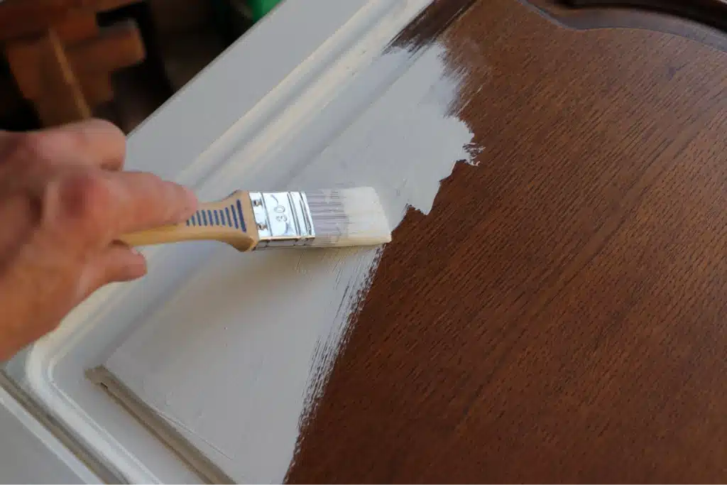 A person's hand using a paintbrush to apply white paint on a wooden surface, partially covering the brown finish. The scene suggests a home improvement or DIY project.