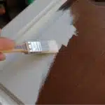 A person's hand using a paintbrush to apply white paint on a wooden surface, partially covering the brown finish. The scene suggests a home improvement or DIY project.