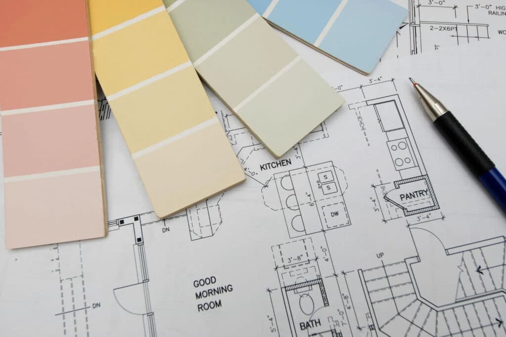 Color swatches in pastel shades are arranged on top of architectural floor plans. A blue pen is placed next to the plans, which include labels for rooms like 'KITCHEN,' 'PANTRY,' and 'BATH.'