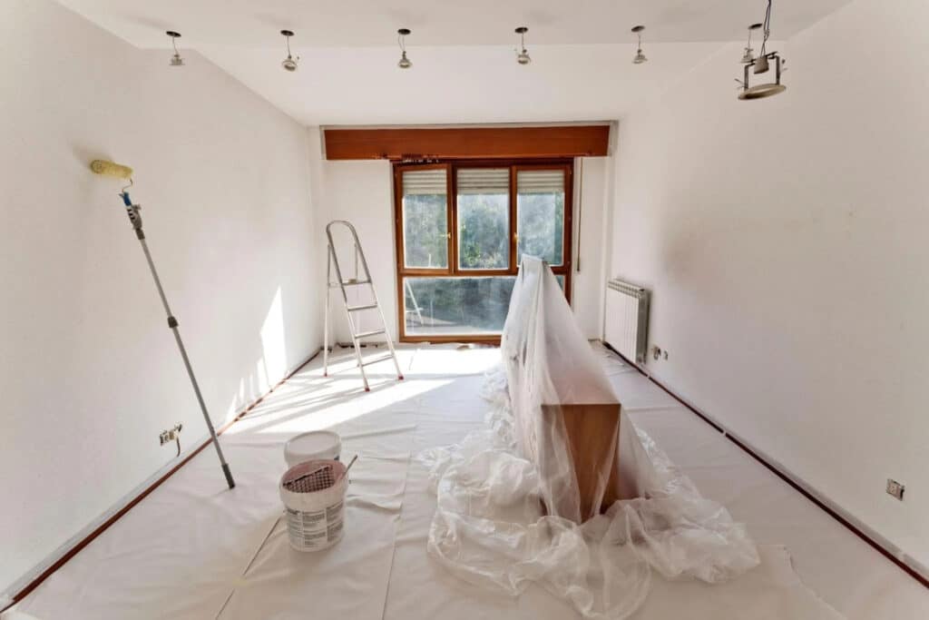 A spacious, empty room with white walls and wooden floors covered with a protective sheet. A ladder, a paint roller on a pole, and a bucket of paint are visible, along with a wooden table draped in plastic, and large windows letting in natural light.