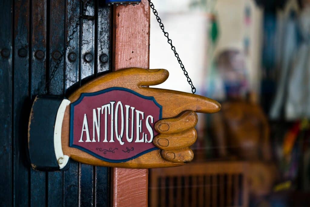 A wooden sign shaped like a hand pointing, featuring the word "ANTIQUE" in white on a red background, hanging from a chain. The background shows blurred interior details of a shop.