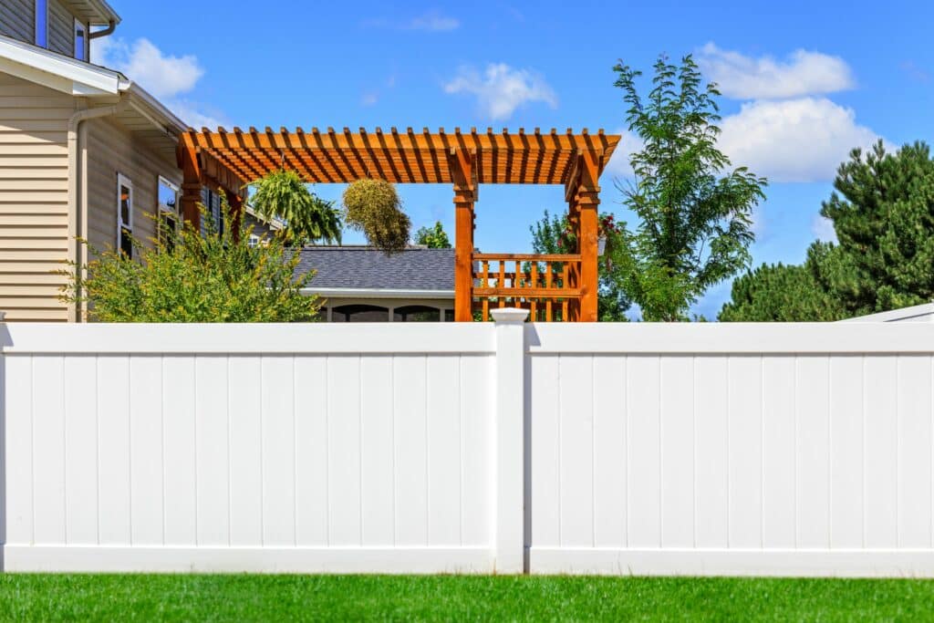 A white vinyl fence surrounding a residential yard, featuring a wooden pergola with plants above. The background shows a clear blue sky and trees, with a well-maintained lawn in the foreground.