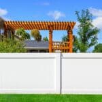 A white vinyl fence surrounding a residential yard, featuring a wooden pergola with plants above. The background shows a clear blue sky and trees, with a well-maintained lawn in the foreground.