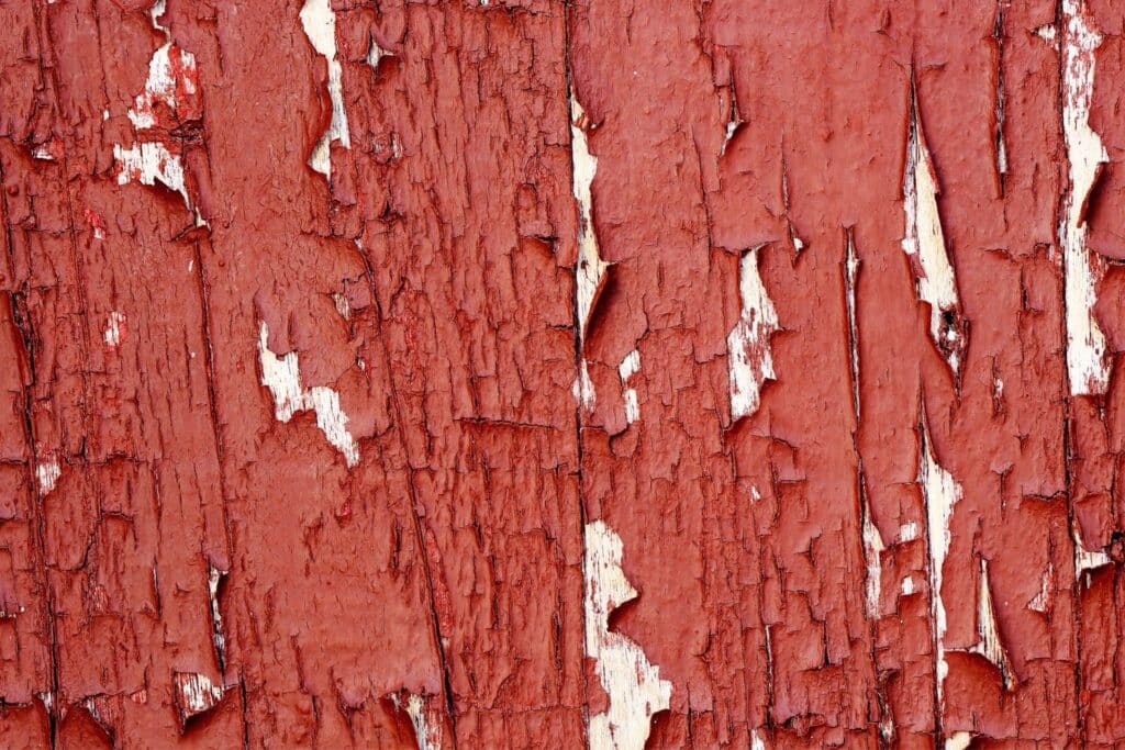 Close-up of weathered red paint on a wooden surface, showing cracks and peeling layers, with visible sections of white underneath.