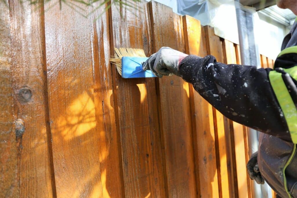 A person applying a blue paint on a wooden fence with a paintbrush. The fence is stained a warm brown color, and sunlight casts shadows on the surface.