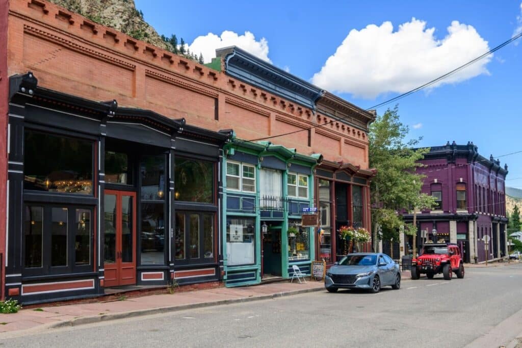 A street view of historical buildings with colorful facades, including a green building and purple structure, along a road. A gray car is parked nearby, with a red vehicle visible further down the street. Blue sky with clouds overhead.