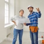 A construction worker in a hard hat discussing plans with a woman in casual clothing, both smiling. The room is partially renovated with tools and a ladder visible in the background.