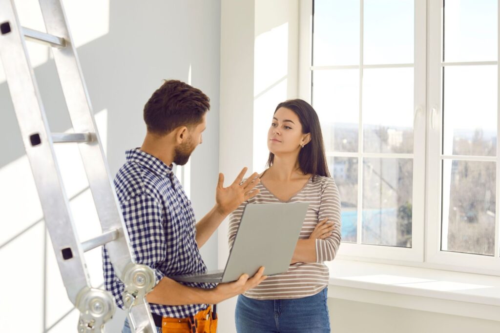 A man and a woman are discussing a project in a bright room with large windows. The man holds a laptop and gestures, while the woman listens attentively. A ladder is visible in the background.